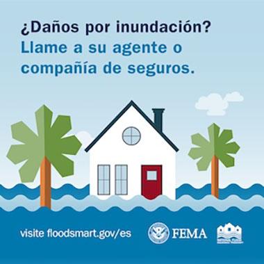 Una casa blanca con una puerta roja está entre dos palmeras, rodeada de agua de inundación bajo un cielo azul con nubes.