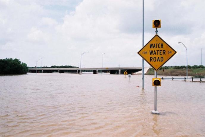 A sign reading "watch for water on road" stands above a flooded road.